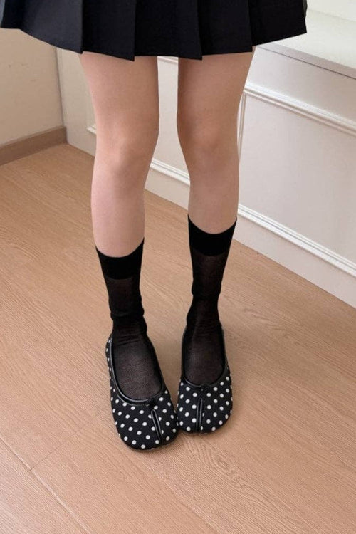 Black polka-dot Mary Jane shoes with black sheer socks, shown on a model's feet in an indoor setting with light wood flooring and white paneled walls.