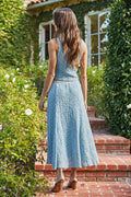 Woman in a light blue dress standing on a brick path with greenery around.