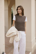Woman wearing a brown knitted vest over a white shirt with white pants, standing outdoors.