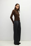 A woman modeling a brown ruched long-sleeve mock neck top paired with dark charcoal wide-leg trousers, standing against a light gray studio background.