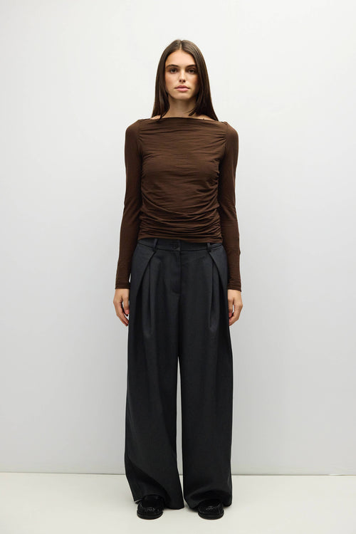 A model stands against a light gray studio background wearing a brown off-the-shoulder long-sleeve top and black wide-leg trousers.