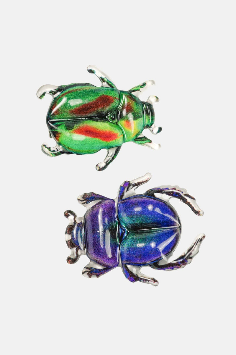 Two iridescent beetle-shaped brooches on a plain light gray background. The top brooch is metallic green with red and yellow shifts, and the bottom brooch is metallic blue and purple with silver-toned legs and antennae.