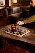A person in a dimly lit living room lights a candle placed on a wooden game board on a rustic table. The candle is in a dark amber-brown glass jar with a small red label, and the board has black-and-white squares with small game pieces.