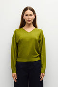 A woman stands against a plain light gray studio wall wearing a loose olive green V-neck sweater, dark navy wide-leg pants, and brown flats.