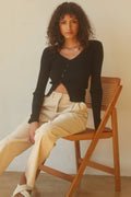 Woman sitting on a wooden chair wearing a black cardigan and beige pants.