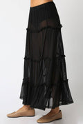 A black tiered maxi skirt with a sheer mesh overlay, shown on a model wearing beige flat shoes against a plain white background.
