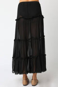 A black tiered sheer maxi skirt with ruffled trim, shown on a model from the waist down wearing beige flat shoes against a plain white background.