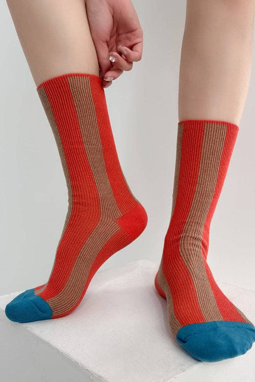 A person wearing a pair of crew socks with a red and beige vertical stripe pattern and blue toes, standing on a white block against a light gray background.