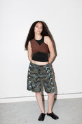 Woman wearing a brown top and camouflage shorts against a white wall.