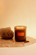 A lit amber glass candle with two flames sits on a stone slab beside a rough stone and a match, against a warm beige background.