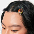 A close-up of a person's dark hair featuring a small, realistic snail hair clip holding one side of the hair. The snail has a glossy, multi-colored shell with orange, yellow, and green tones.