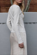 Woman in a white long-sleeve top and polka dot skirt standing against a gray wall with cacti in the background.