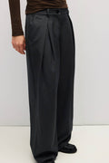 A person wearing dark charcoal wide-leg pleated trousers, styled with a brown long-sleeve top and black loafers, photographed against a plain light gray studio background.