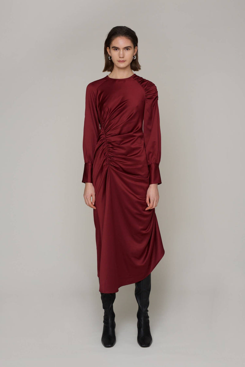 A woman modeling a long-sleeved, gathered midi dress in deep burgundy with black boots, standing against a neutral light gray studio background.