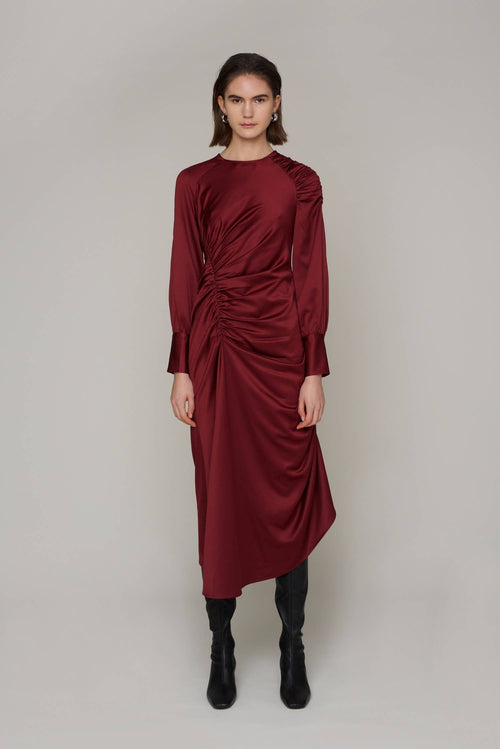 A woman modeling a long-sleeved, gathered midi dress in deep burgundy with black boots, standing against a neutral light gray studio background.