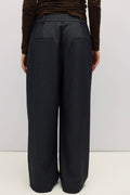 A person shown from the back wearing dark charcoal wide-leg trousers and a brown long-sleeve top, standing against a plain light gray studio background.