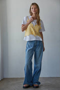 Woman wearing a white t-shirt with a yellow floral crochet top over it against a plain background