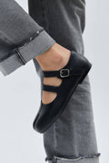 Black flat shoe with a buckled strap worn by a person in gray pants on a light gray background