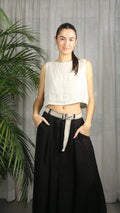Woman wearing a white sleeveless top and black wide-leg pants standing against a gray curtain.