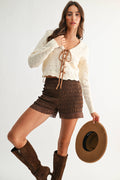 Woman wearing a white lace top, brown shorts, and boots, holding a hat on a plain background