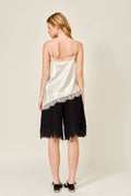 Woman wearing a cream satin camisole with lace trim and black shorts on a plain background