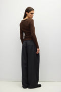 A woman stands in profile wearing a fitted brown long-sleeve top and very wide-leg black pants against a plain light gray studio background.