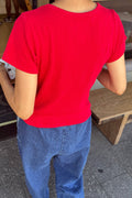 A person viewed from behind wearing a bright red short-sleeve crewneck t-shirt and blue jeans, standing in an outdoor or semi-outdoor setting.