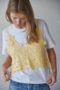 Woman wearing a white t-shirt with a yellow floral crochet top over it against a plain background