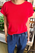 A person wearing a bright red short-sleeve crewneck t-shirt and blue jeans, standing in front of a store display with bottles.