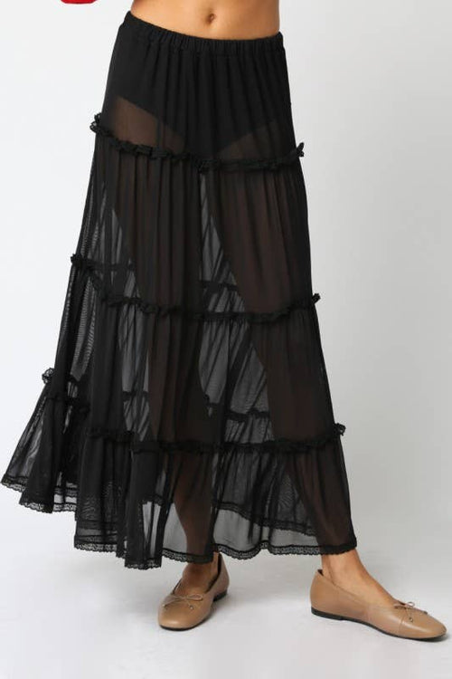 A black tiered sheer maxi skirt with ruffled trim, shown on a model wearing beige flat shoes against a light gray studio background.