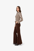 A woman modeling a retro-style brown and beige vertically striped long-sleeve blouse with ruffled cuffs, paired with dark brown flared trousers and black pointed-toe heels, set against a plain light gray studio background.