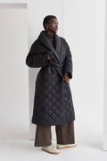 A model wearing a black quilted puffer wrap coat with a folded front collar and a self-belt with loops on the side. The coat has hidden pockets and is oversized for comfort.
