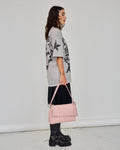 A woman stands in profile wearing an oversized light gray graphic T-shirt, a dark pleated skirt, black platform boots, and holding a pink studded shoulder bag against a plain light gray studio background.