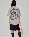 A woman shown from the back wearing a light gray oversized graphic T-shirt with a large black gothic-style print, a black pleated skirt, and black knee-high boots, while holding a pink studded shoulder bag against a plain light gray studio background.