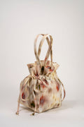 A small drawstring bag with a soft, painterly abstract print in beige, cream, muted red, and olive green, featuring two long handles, photographed on a seamless light gray studio background.