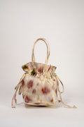 A small drawstring bucket bag with a beige, cream, and white base color and abstract red, green, and brown brushstroke patterns, featuring double top handles, photographed on a light gray background.