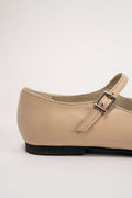 A close-up side view of a beige leather Mary Jane flat shoe with a silver buckle strap and a black sole, set against a plain light gray background.