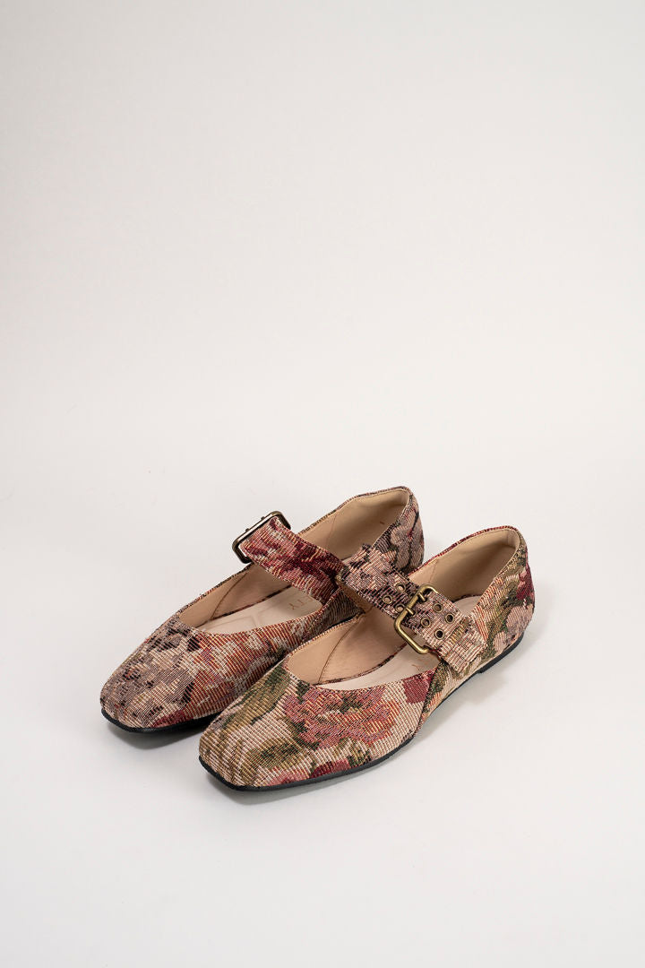 A pair of Mary Jane flats with a woven floral pattern in muted pink, brown, and green, featuring a single strap and a gold-tone buckle, shown on a light gray background.