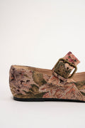 A close-up side view of a flat shoe with a woven multicolor pattern in shades of brown, beige, and red, featuring a brown strap and an antique bronze buckle.
