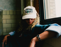 A person wearing a cream-colored baseball cap with a 'LOVE' graphic, sitting by a window in a dimly lit room.