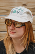 A woman wearing a white trucker-style cap with 'IMAGINE A WORLD' and a spiral graphic on the front, along with tortoiseshell sunglasses, shown against a wooden background.