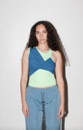 A model with long curly dark hair stands against a plain light gray background wearing a color-blocked sleeveless top with blue, navy, and pale yellow-green panels, paired with light blue pants.