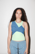 A model with long, curly dark hair is wearing a color-blocked athletic tank top in blue and pale yellow-green, paired with light blue pants, against a plain off-white background.