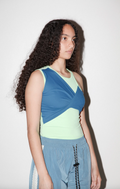 A model with long, curly dark hair is shown from the waist up, wearing a color-blocked athletic tank top in blue, mint green, and light yellow, paired with light blue pants against a plain white background.