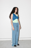 A model stands against a plain white wall wearing a color-blocked sleeveless top with blue, lime green, and navy panels, paired with wide-leg light blue jeans featuring a decorative hanging chain detail on the side.