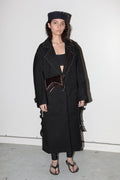 Person wearing a long black coat with studded details and a hat against a white background