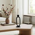 Decorative black candle holder with a white candle on a wooden table, next to a vase with dried flowers and a book.