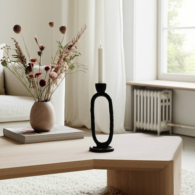 Decorative black candle holder with a white candle on a wooden table, next to a vase with dried flowers and a book.