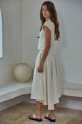 Woman wearing a white outfit standing in a minimalistic room with a vase on a ledge.