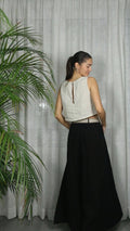 Woman wearing a white sleeveless top and black skirt standing against a light curtain with a plant to the left.